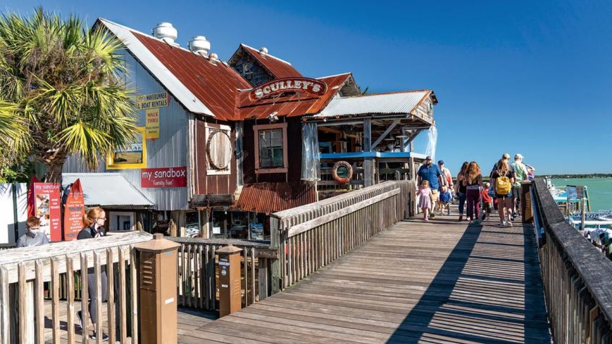 John’s Pass Village & Boardwalk John's Pass Village Boardwalk with visitors walking, featuring Sculley's restaurant and surrounding palm trees, highlighting a vibrant shopping and dining destination in Treasure Island.