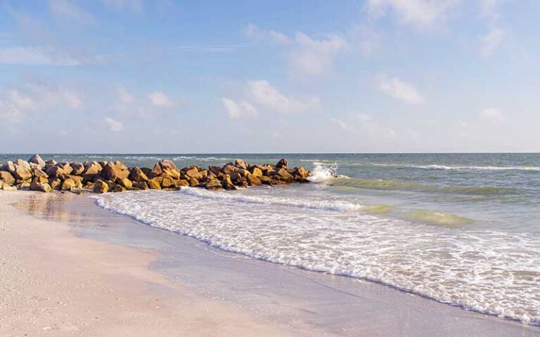 Sunset Beach-min Scenic view of Sunset Beach with gentle waves lapping against rocky shoreline under a blue sky, highlighting the tranquil atmosphere ideal for relaxation and exploration in Treasure Island.